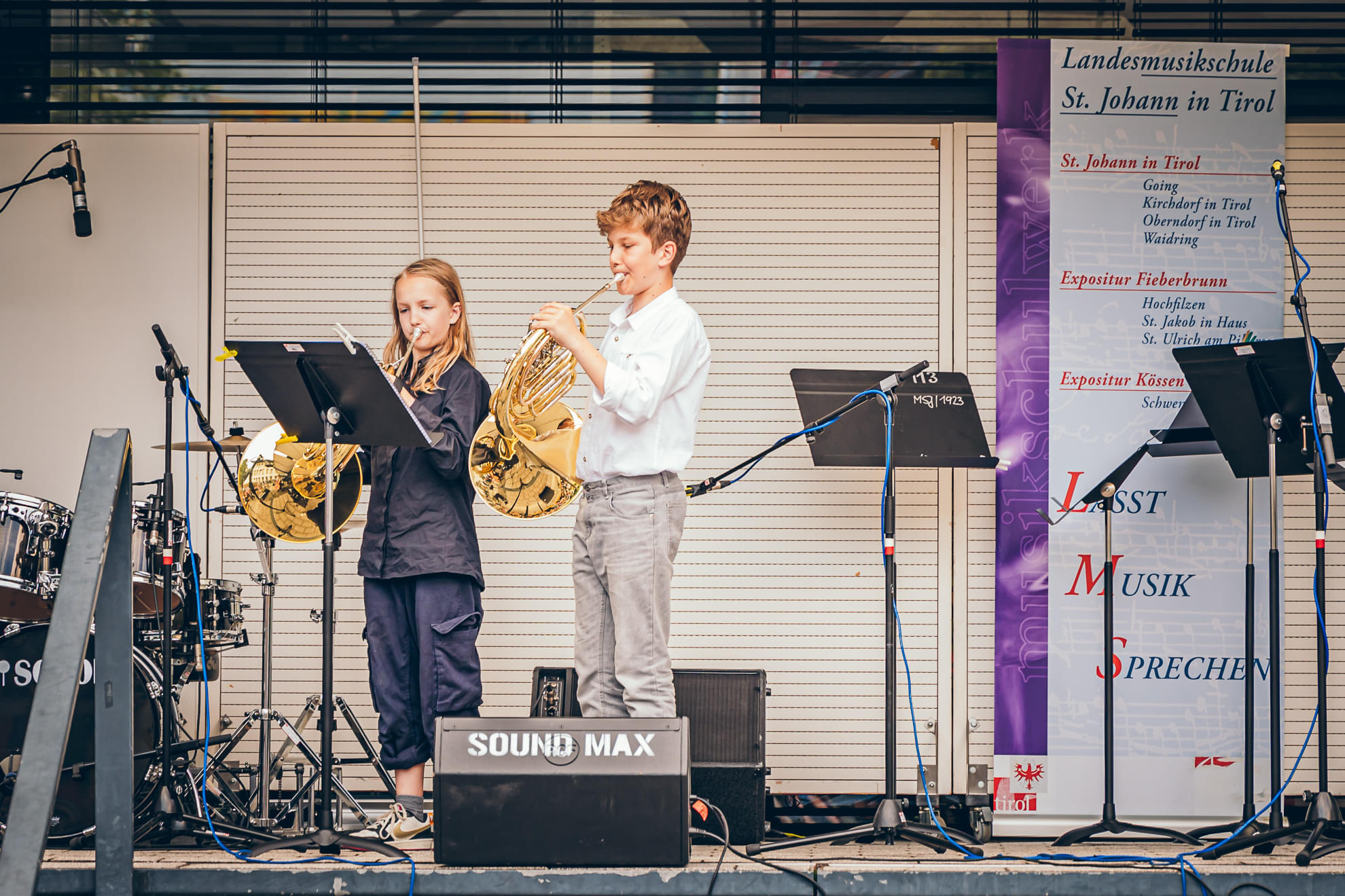 Zwei junge Kinder spielen auf einer Bühne Horn, umgeben von Mikrofonen und Schlagzeug, vor einem Banner, das eine Musikschule bewirbt.
