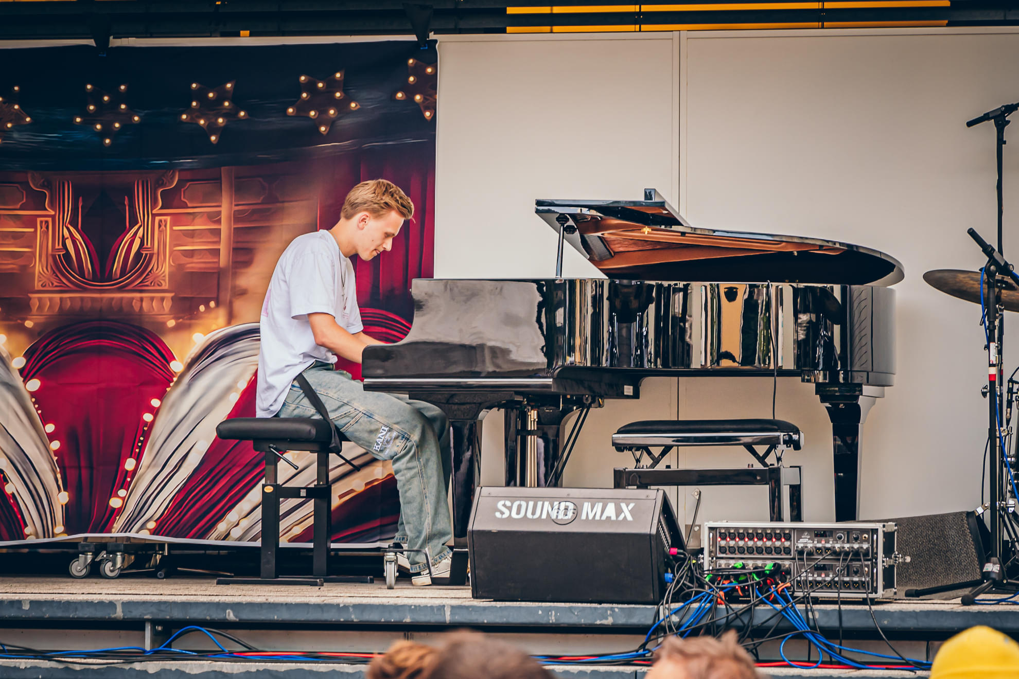 Ein junger Mann spielt Klavier auf einer Bühne mit einem geschmückten Hintergrund. Neben dem Klavier stehen Tonverstärker und Kabel.