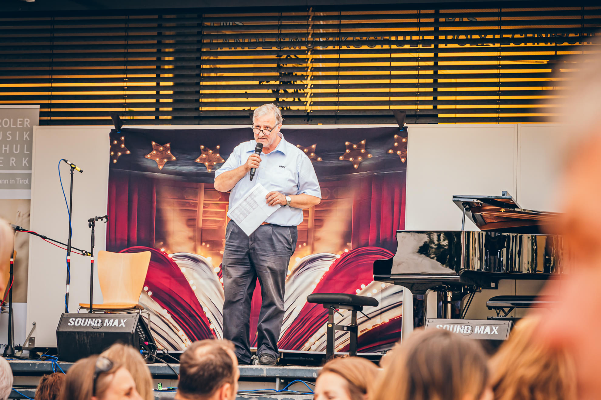 Ein Mann steht auf einer Bühne und spricht in ein Mikrofon. Im Hintergrund sind geschlossene rote Vorhänge und ein Flügel zu sehen. Publikum im Vordergrund.