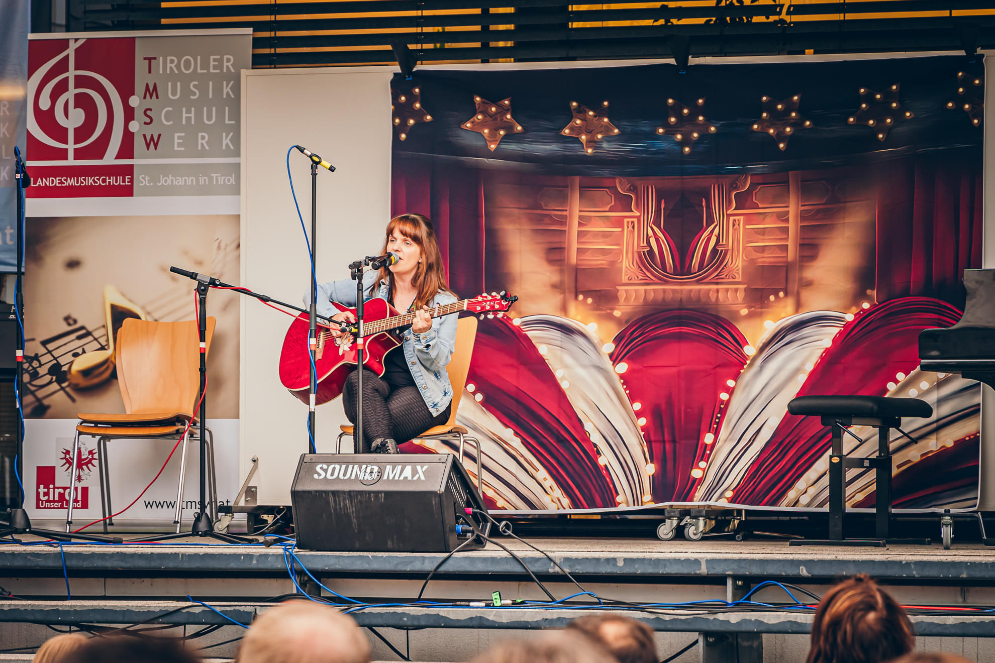 Frau auf Bühne spielt Gitarre bei einem Musikfestival, umgeben von Mikrofonen und Lautsprechern vor einem kunstvollen Hintergrund.