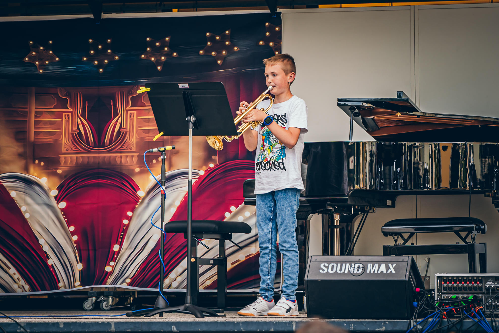 Ein Junge spielt Trompete vor einem Mikrofon auf einer Bühne, neben ihm steht ein Klavier. Der Hintergrund zeigt bunte Beleuchtung und Dekorationen.