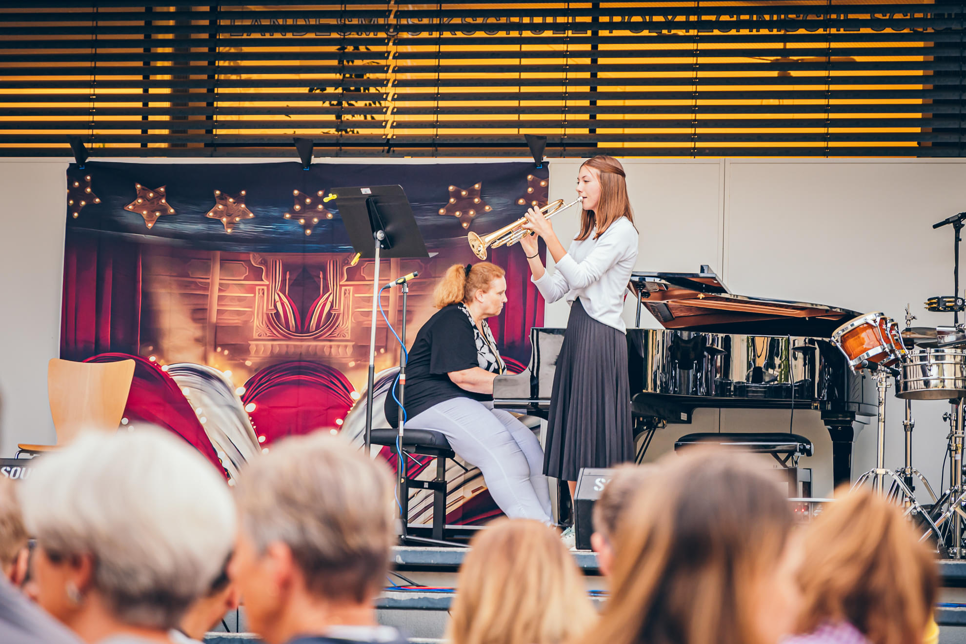 Ein junges Mädchen spielt Trompete auf einer Bühne, eine andere Person sitzt daneben, während Publikum in einem Theater draußen zuschaut.