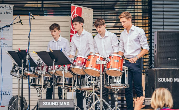 Vier junge Männer in weißen Hemden spielen im Freien auf einer Reihe von Trommeln und Notenständern bei einem Musikauftritt.