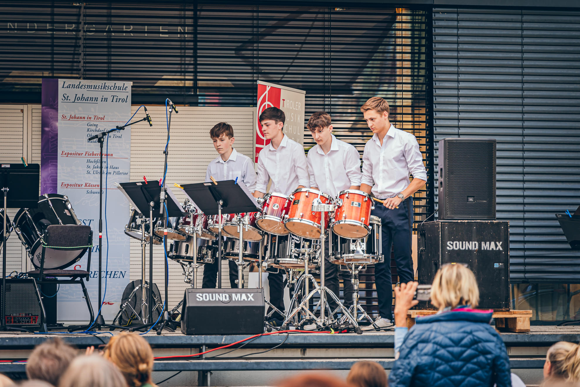 Vier junge Männer in weißen Hemden spielen Schlagzeuge auf einer Bühne vor einem sitzenden Publikum. Im Hintergrund sind Lautsprecher und ein Banner sichtbar.
