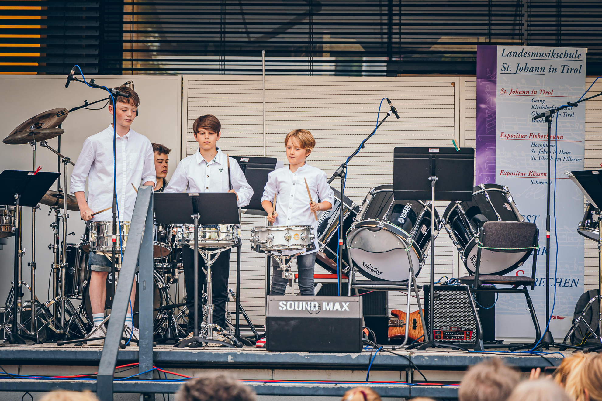 Fünf junge Percussionisten in weißer Kleidung stehen auf einer Bühne mit verschiedenen Schlaginstrumenten, umgeben von Notenständern und Mikrofonen.