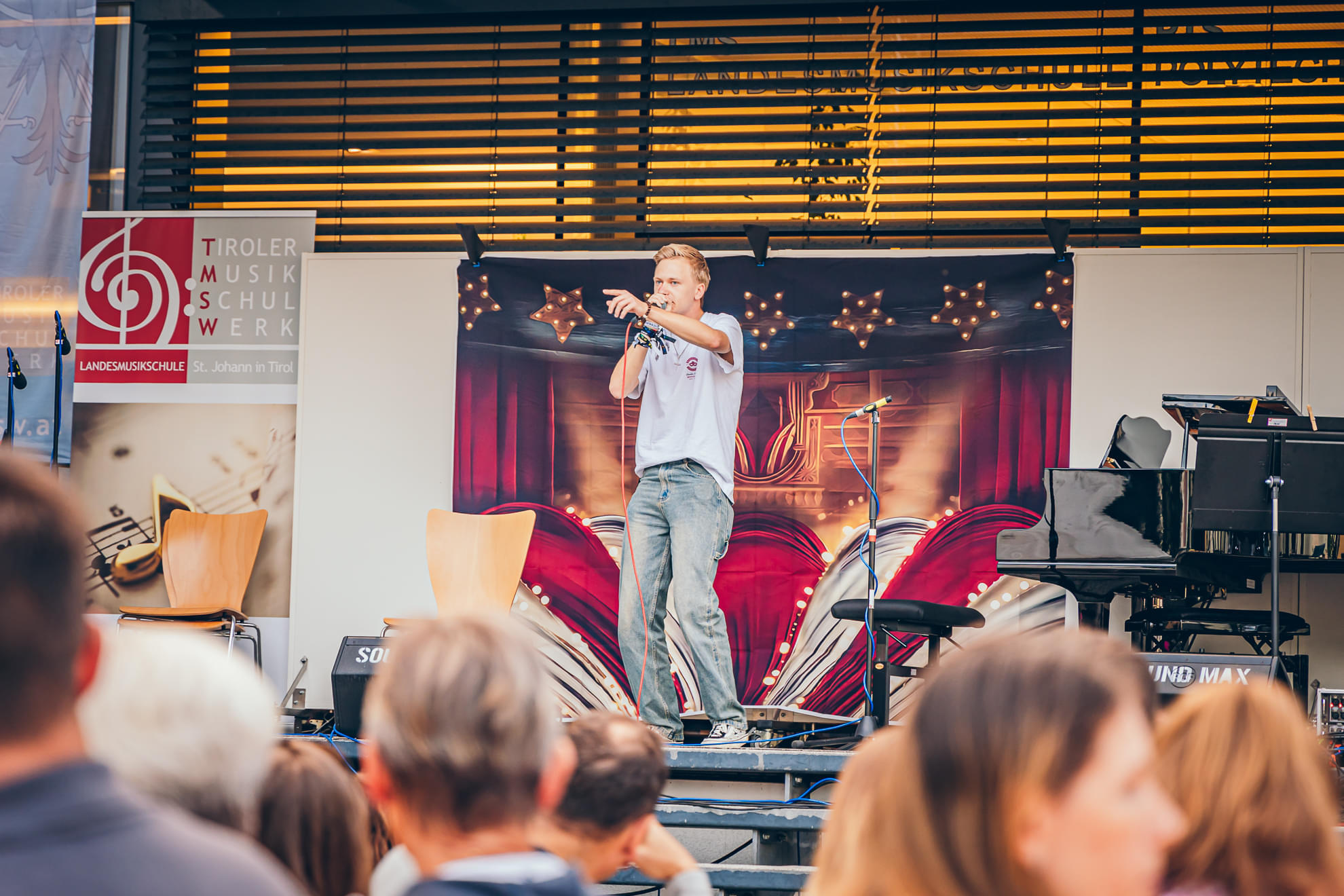Ein Mann hält eine Rede auf einer Bühne im Freien. Hinter ihm ein Klavier und ein großes Plakat. Zuschauer hören gespannt zu. Sommerliche Atmosphäre.