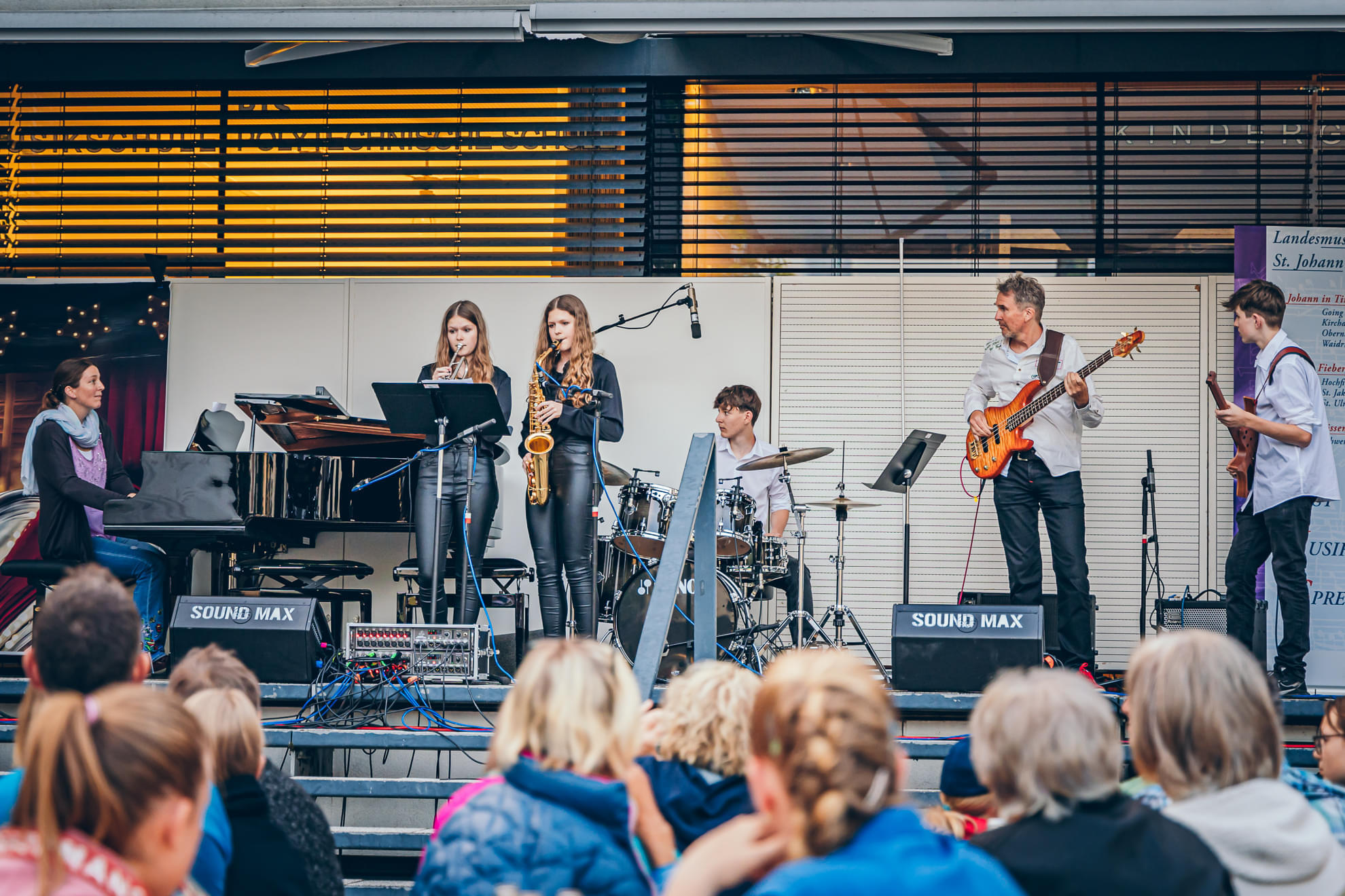 Bandmitglieder spielen auf einer Bühne vor Publikum, darunter ein Saxophonist, ein Keyboardspieler und ein Schlagzeuger. Die Atmosphäre wirkt lebendig und musikalisch.
