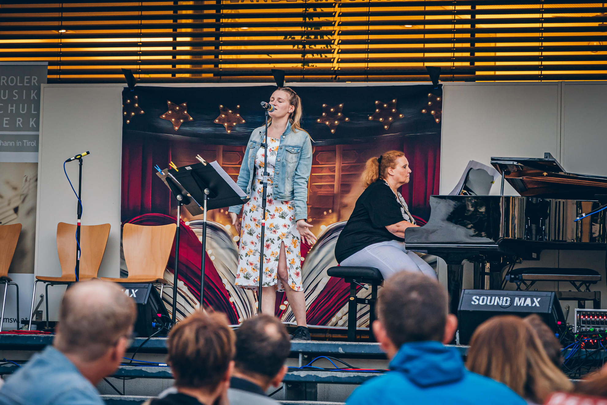Eine Frau singt auf einer Bühne, begleitet von einer Frau am Klavier. Einige Zuschauer sitzen im Vordergrund. Im Hintergrund sind Lichter und Dekorationen.