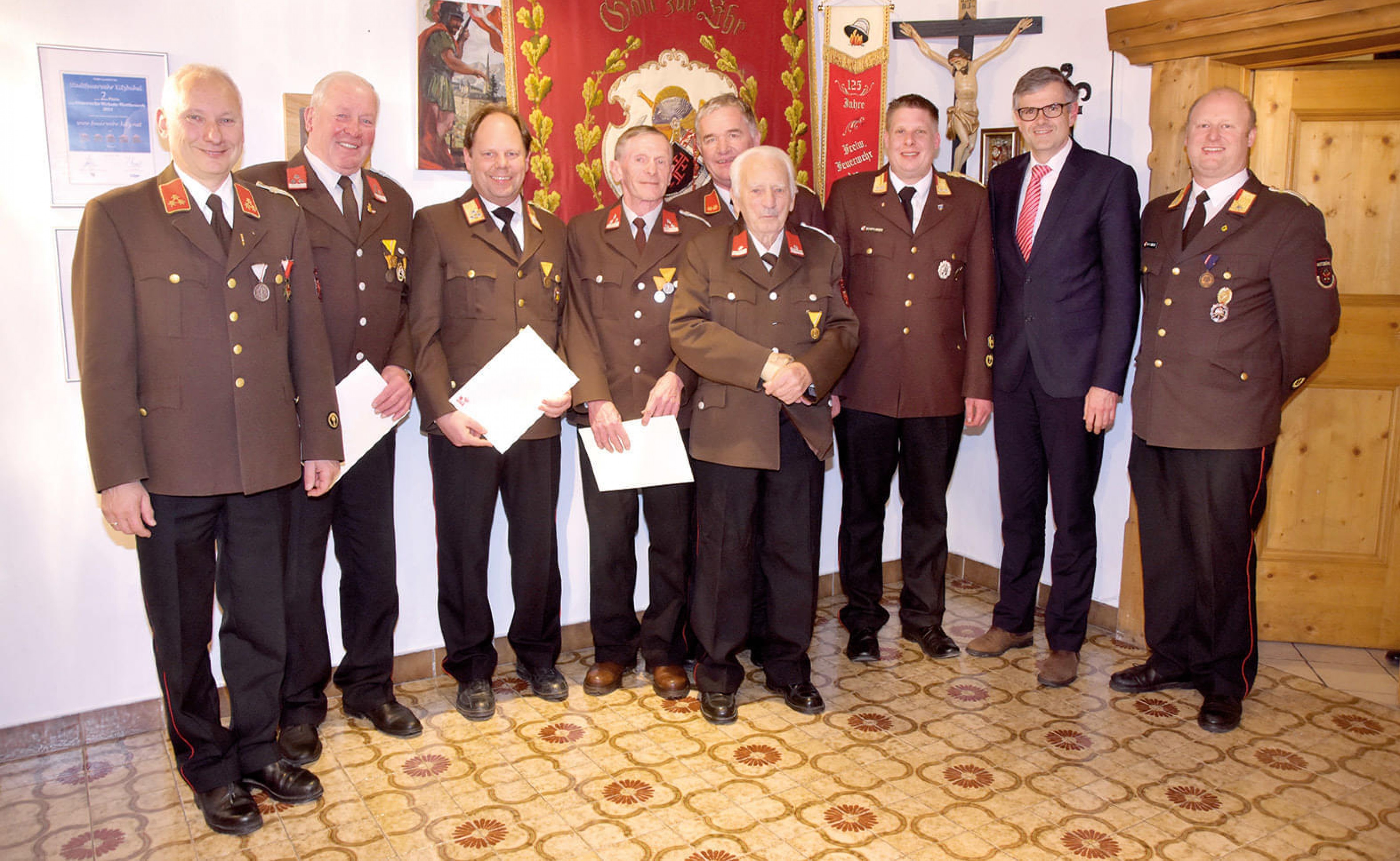 Eine Gruppe von Männern in Uniformen, wahrscheinlich Mitglieder einer Feuerwehr, posiert lächelnd für ein Gruppenfoto in einem dekorierten Raum.