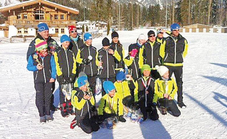 Eine Gruppe von Menschen in Winterkleidung posiert draußen im Schnee vor einem Holzgebäude. Sie tragen bunte Mützen und halten Pokale in den Händen.
