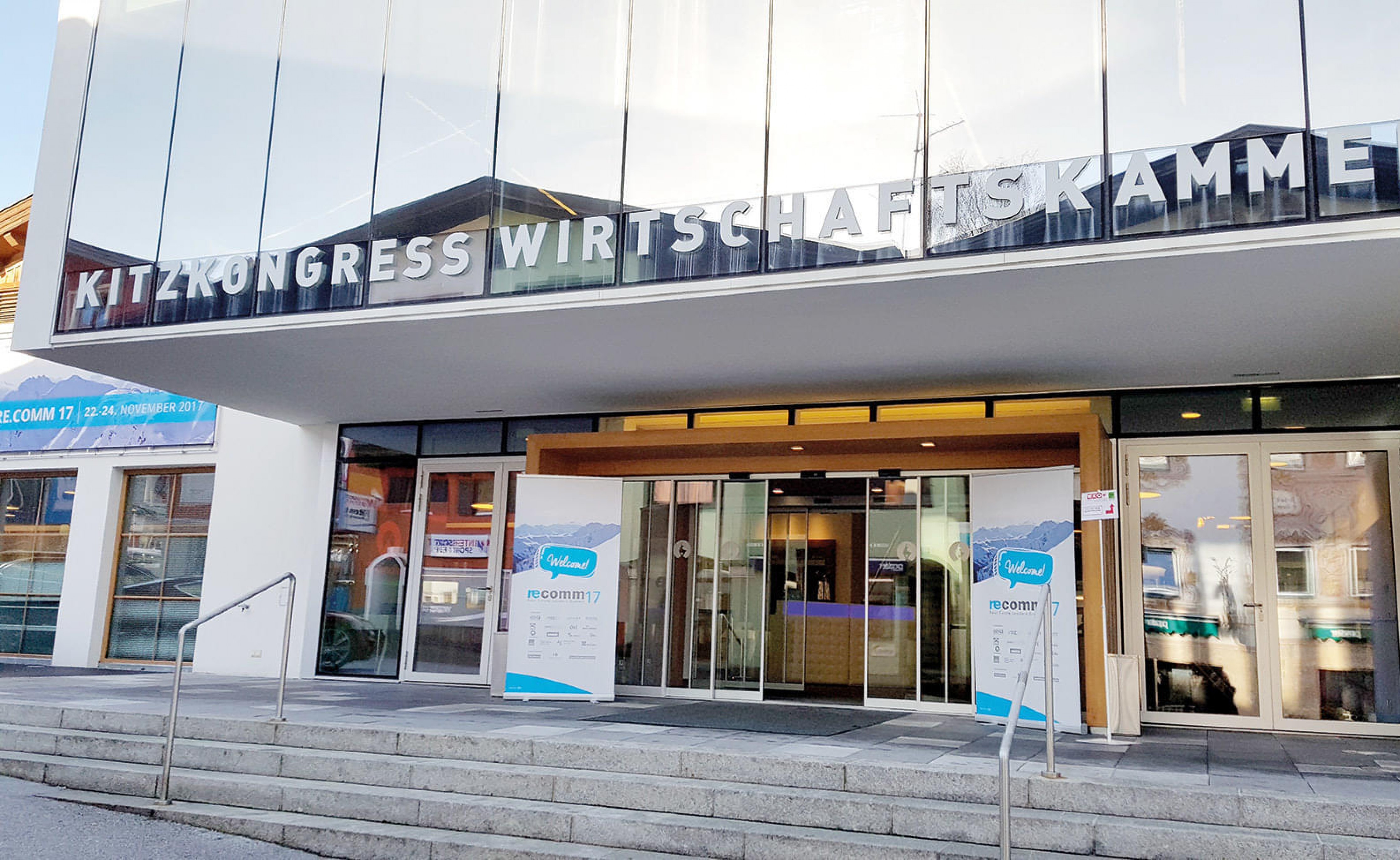 Eingang eines modernen Gebäudes mit der Aufschrift "KitzKongress Wirtschaftskammer" über der Tür. Werbeschilder stehen links und rechts des Eingangs.