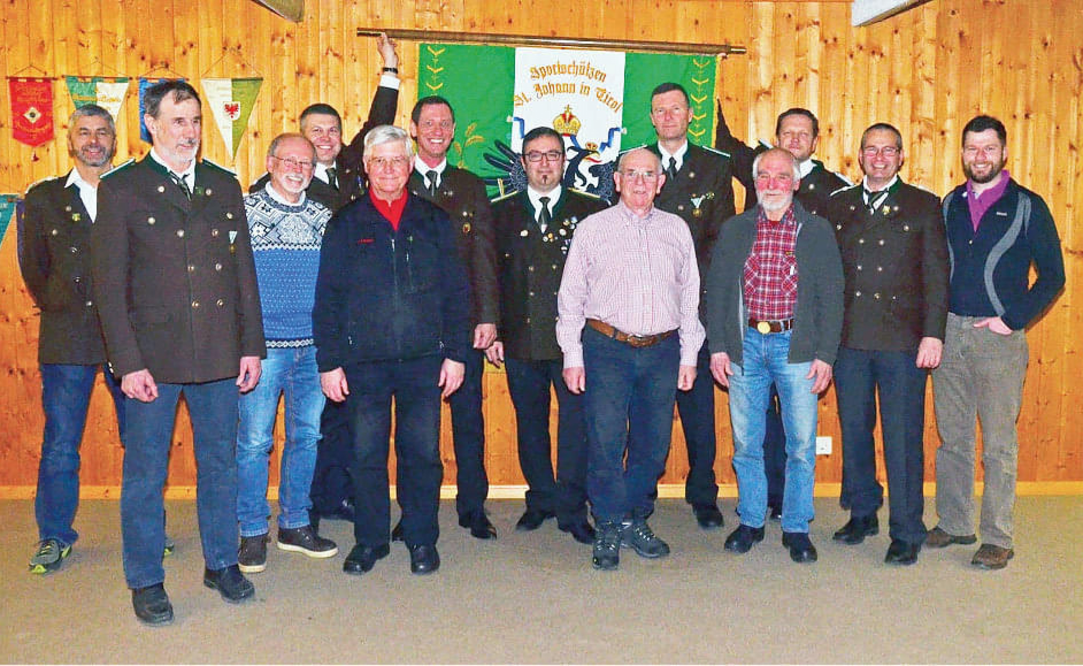Eine Gruppe von Männern, teils in Uniformen, steht vor einer Holzwand mit einem grünen Banner. Sie lächeln und blicken freundlich in die Kamera.