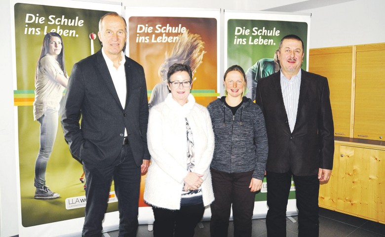 Vier Personen stehen lächelnd vor bunten Bannern mit der Aufschrift "Die Schule ins Leben" in einem Raum mit Holzwänden.