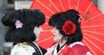 Zwei Personen in traditionellen japanischen Kimonos stehen sich gegenüber, ein roter Regenschirm im Hintergrund, mit Blumen im Haar. Verträumte Szene.