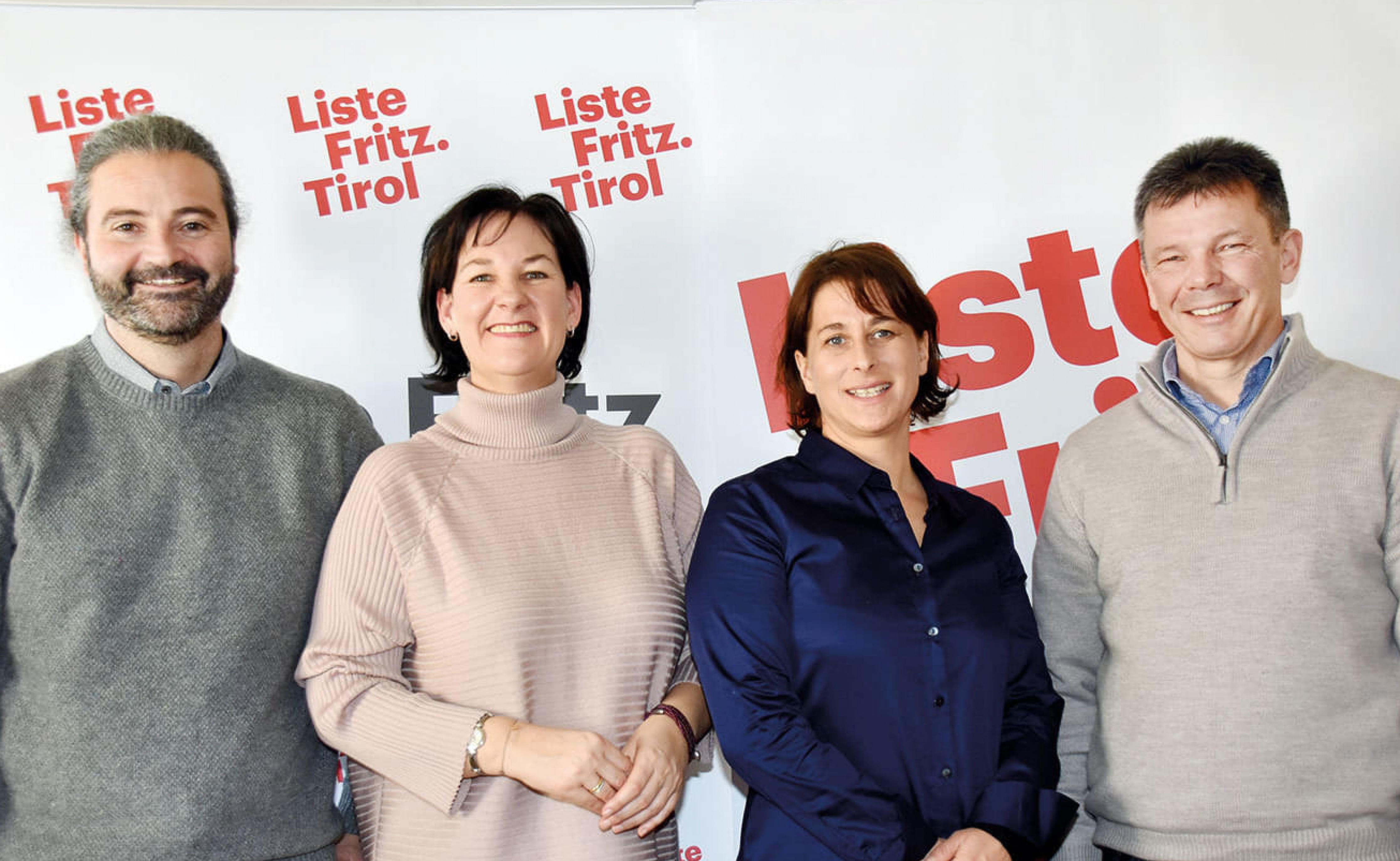 Vier Personen stehen vor einem Banner mit der Aufschrift 'Liste Fritz. Tirol'. Sie wirken fröhlich und freundlich, in Casual-Kleidung.