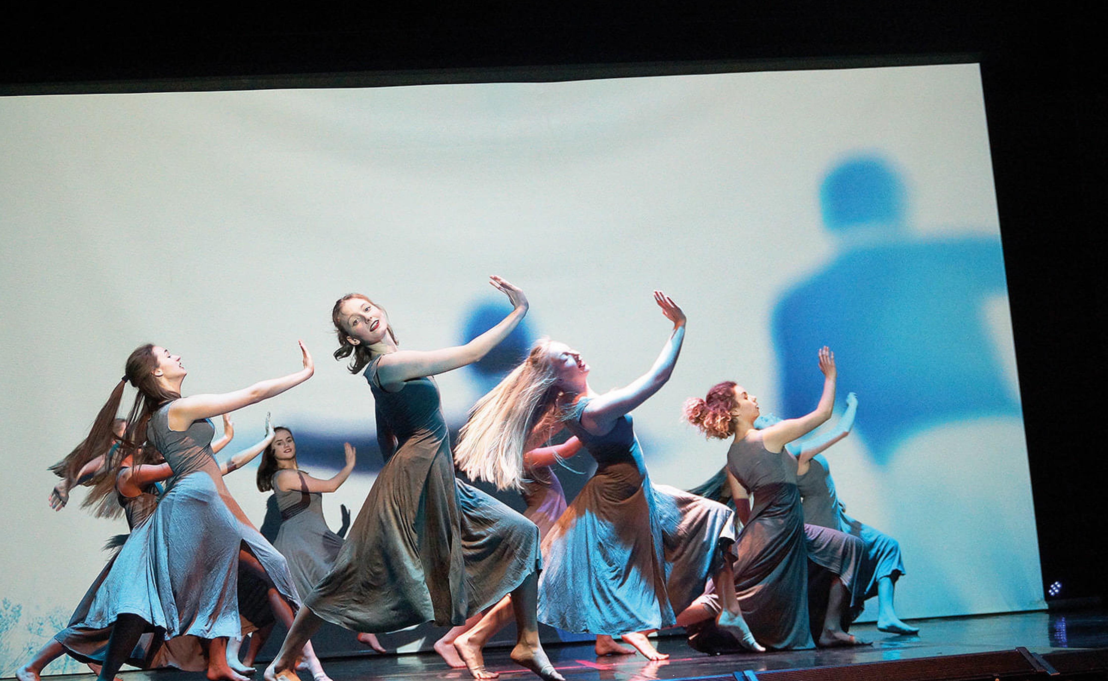 Gruppe von Tänzerinnen in grauen Kleidern auf einer Bühne, die dynamische Bewegungen ausführen. Hintergrund mit blauen, verschwommenen Formen gestaltet.