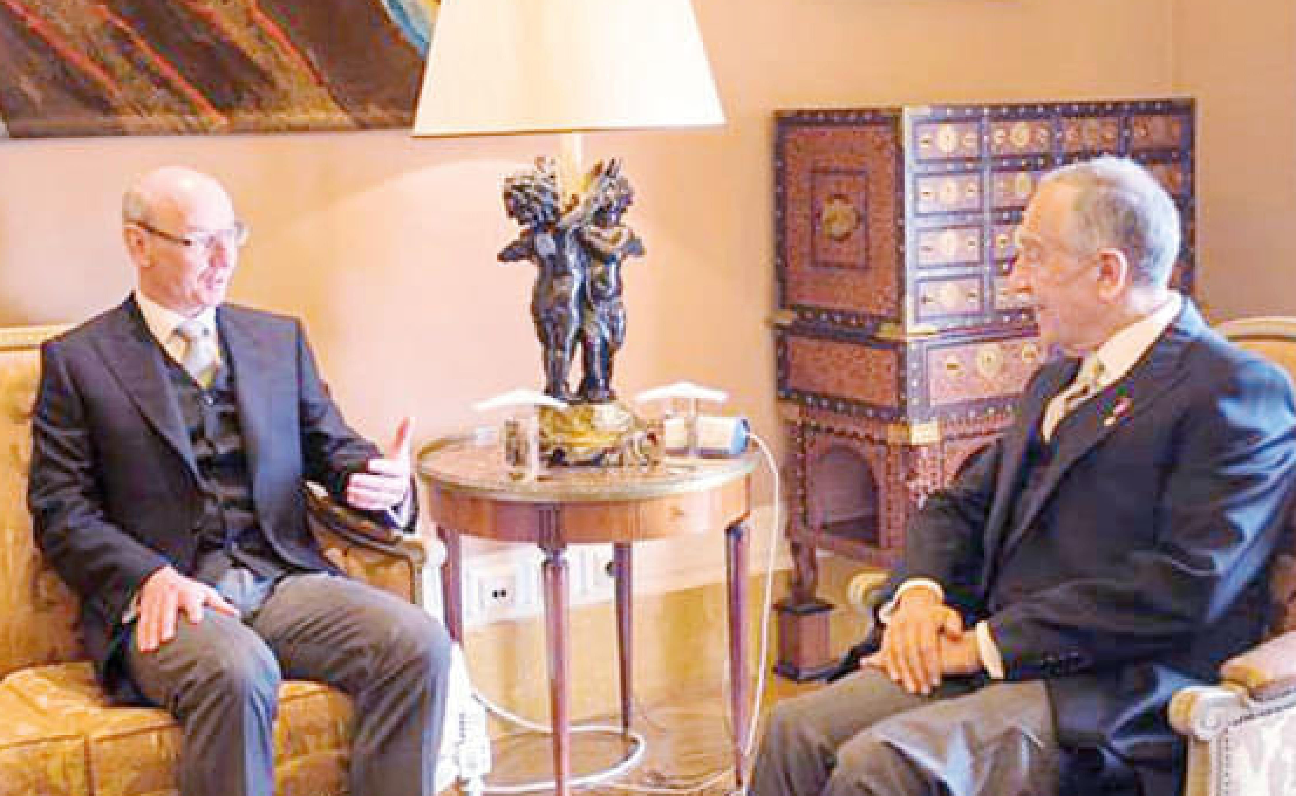Zwei ältere Männer in Anzügen sitzen sich in einem elegant eingerichteten Raum gegenüber und unterhalten sich. Ein kunstvoller Tisch und eine Lampe sind zu sehen.