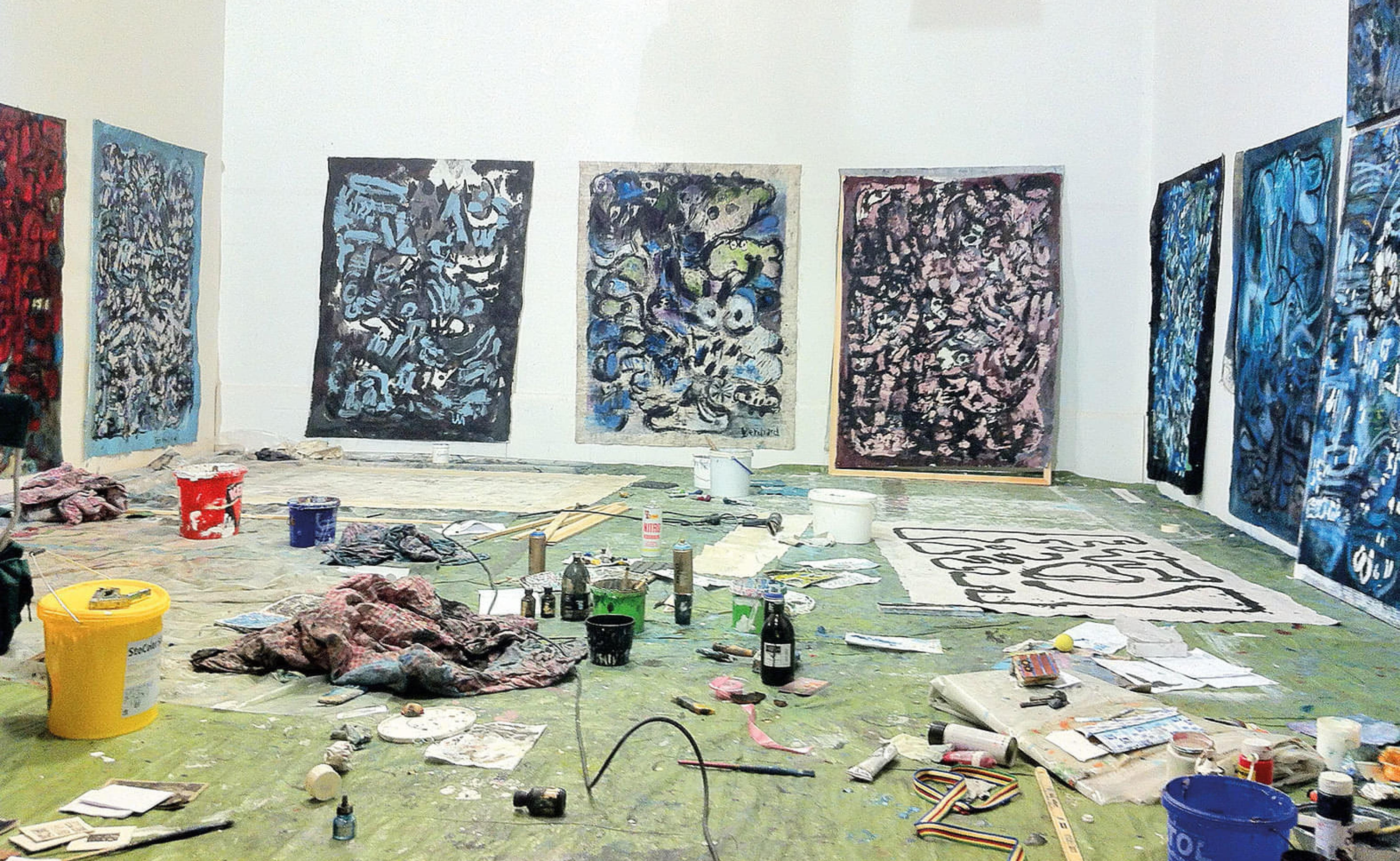 Ein Atelier mit großen abstrakten Gemälden an den Wänden und Malutensilien auf dem Boden. Farben und Leinwände sind verstreut, ein kreatives Chaos herrscht.