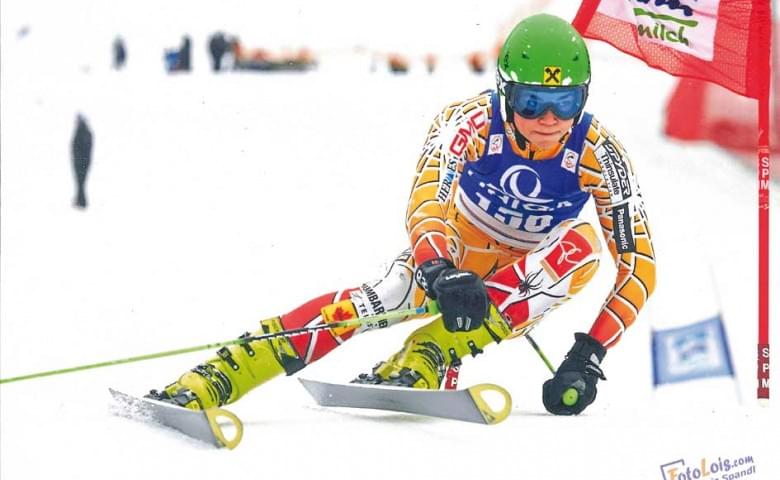 Ein Skifahrer in bunter Kleidung fährt dynamisch durch einen Slalomkurs im Schnee, einen grünen Helm tragend. Schnee wirbelt um ihn herum auf.