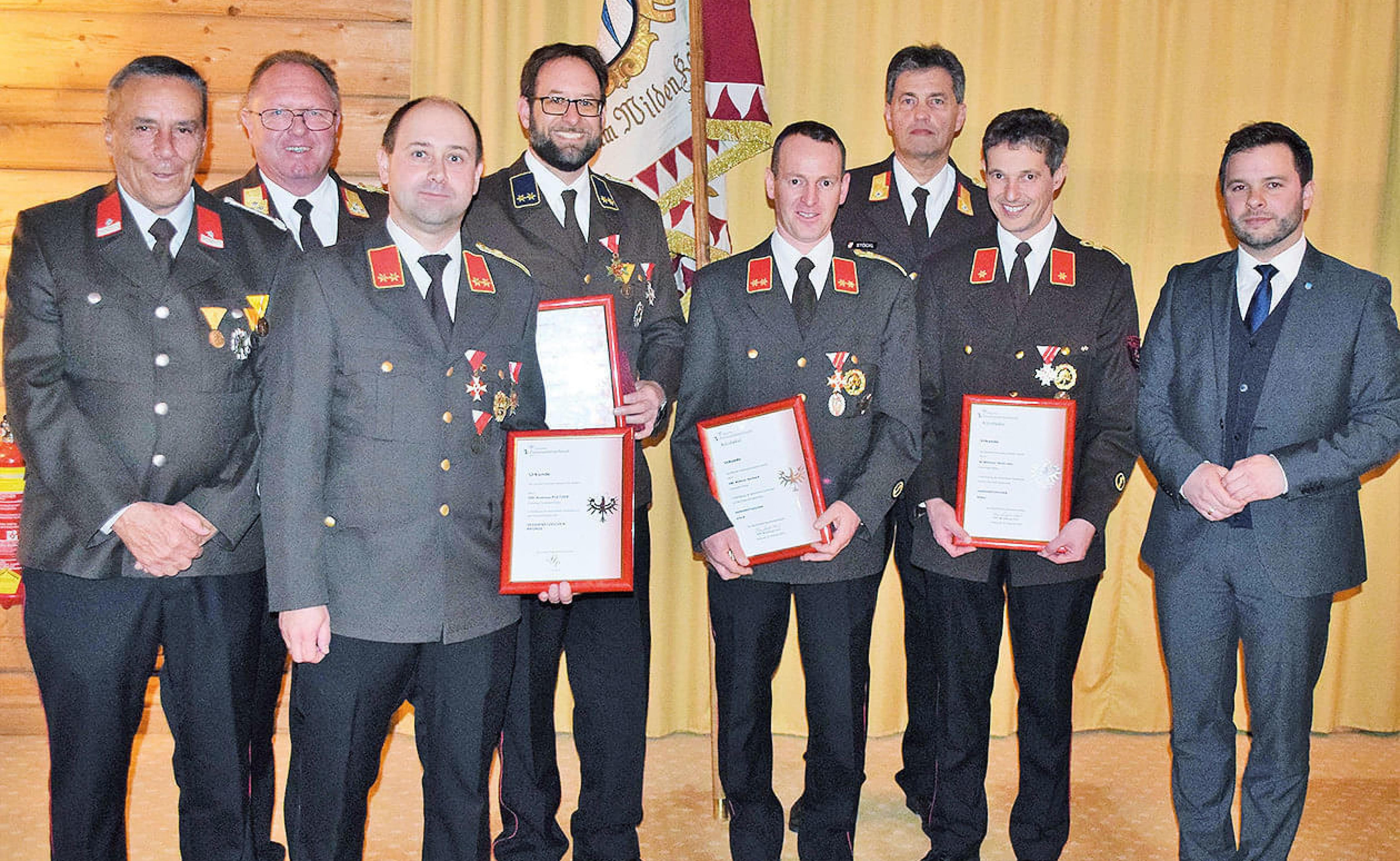 Männer in Uniformen und Anzügen halten Zertifikate. Sie stehen stolz in einer Reihe vor einem gelben Vorhang, einige tragen Abzeichen und Auszeichnungen.