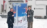 Zwei Männer stehen im Schnee vor einem Rathaus und halten ein Plakat mit winterlichen Motiven. Beide lächeln und zeigen den Daumen nach oben.