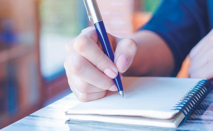 Eine Hand hält einen blauen Stift über ein leeres Notizbuch auf einem Tisch. Sonnenlicht fällt durch ein Fenster und erhellt die Szene.
