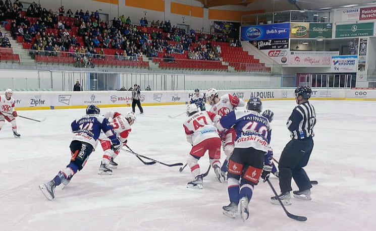 Ein intensives Eishockeyspiel in einer Halle, bei dem Spieler in roten und blauen Trikots um den Puck kämpfen. Zuschauer verfolgen das Spiel von den Rängen.