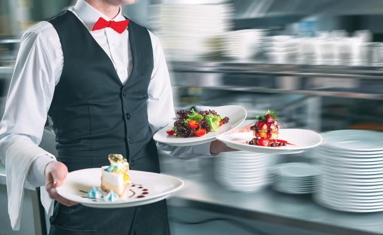 Kellner in schwarzer Weste und roter Fliege trägt drei Teller mit raffinierten Gerichten durch ein geschäftiges Restaurant oder eine Küche.