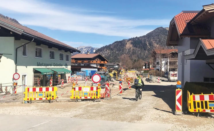 Straßenbaustelle in einer ländlichen Umgebung mit Bergen im Hintergrund. Verkehrsschilder und Absperrungen lenken Fahrzeuge um. Ein Radfahrer fährt vorbei.