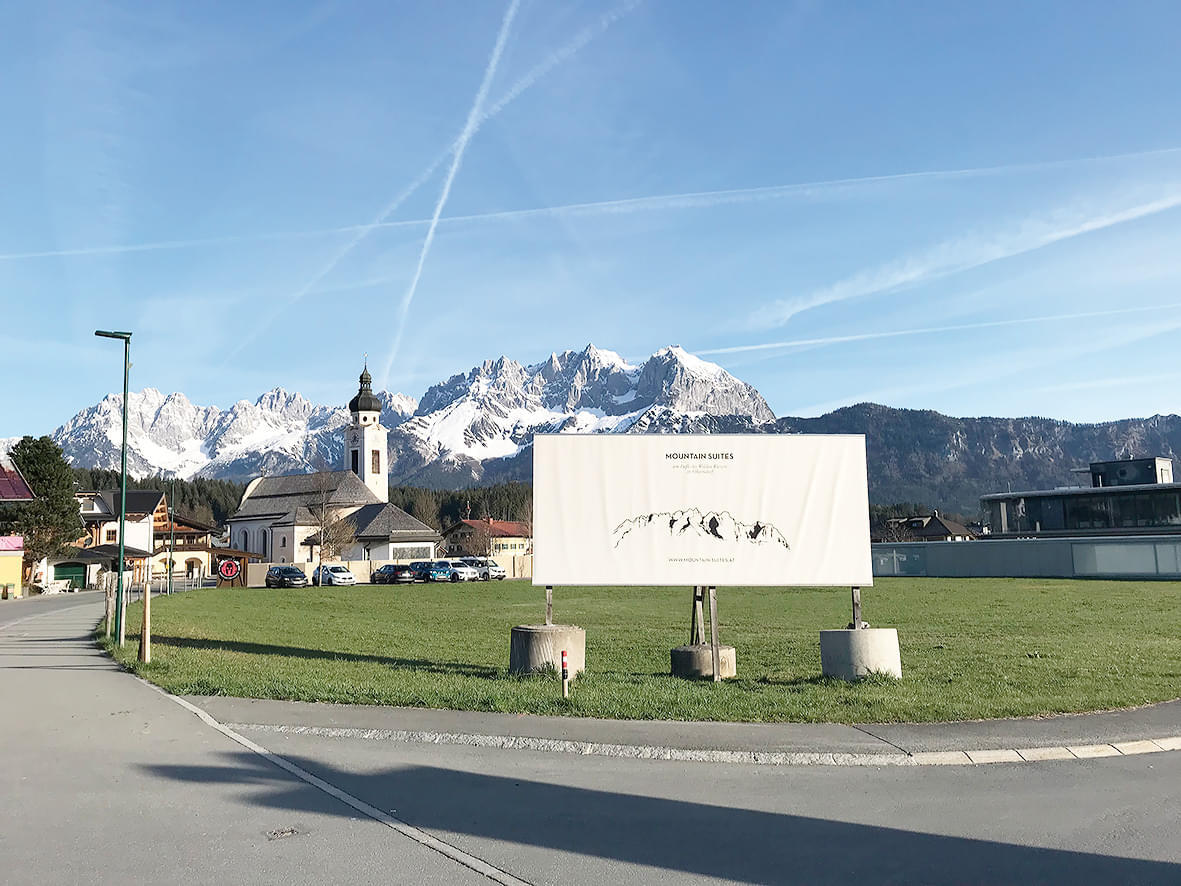Eine Kirche vor schneebedeckten Bergen und einem klaren blauen Himmel mit Kondensstreifen, im Vordergrund eine Werbetafel auf einer Wiese.