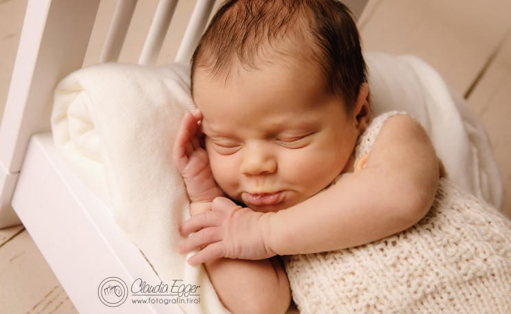 Schlafendes Baby auf einer weichen Decke, den Kopf sanft mit der Hand gestützt, in einem cremefarbenen Strickoutfit, auf hellem Hintergrund.