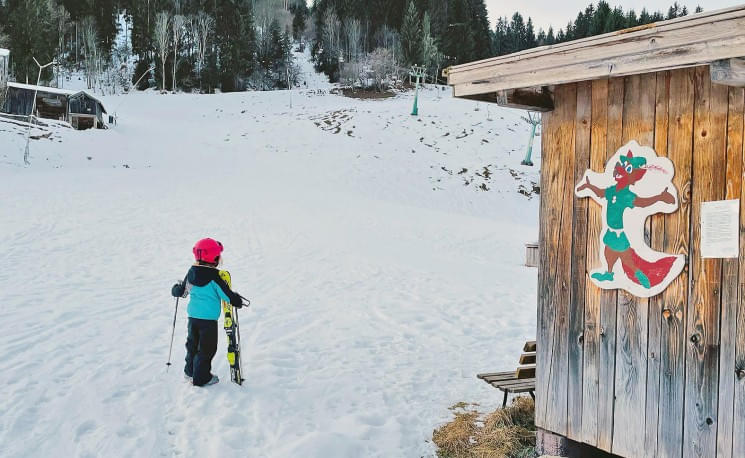 Ein Kind in bunter Winterkleidung steht mit Skiern im Schnee vor einer Holzhütte mit einem lustigen Tierbild. Im Hintergrund sind Bäume und eine Skipiste.