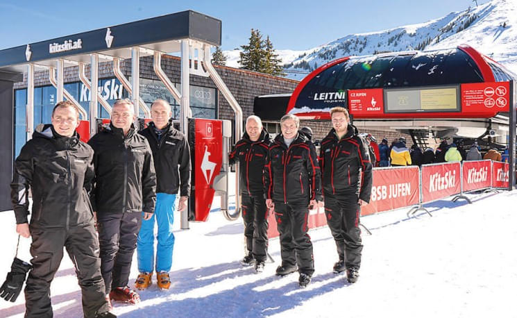 Gruppe von Menschen in Skibekleidung steht vor einem Ski-Lift bei sonnigem Winterwetter. Im Hintergrund sind schneebedeckte Berge und klare blaue Himmel.