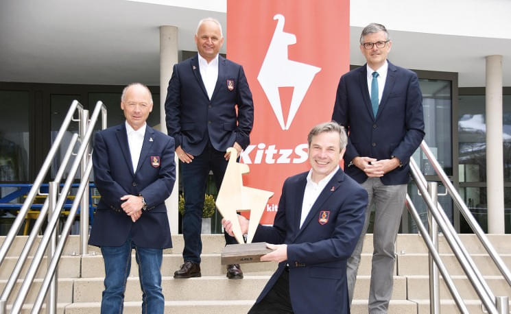 Vier Männer in Anzügen stehen auf einer Treppe vor einem Gebäude. Einer hält eine kleine Skulptur. Im Hintergrund ist ein rotes Banner mit Schriftzug sichtbar.