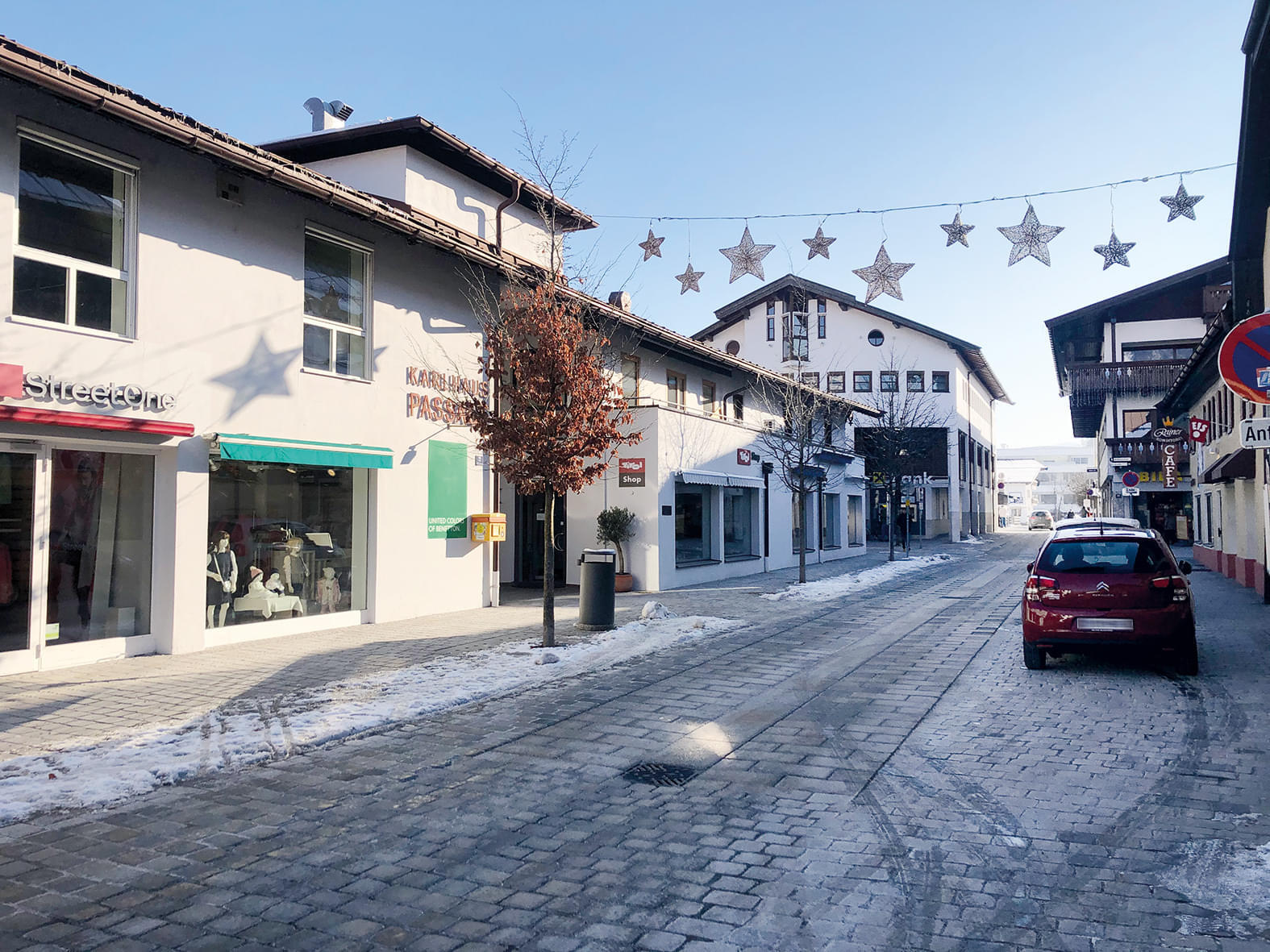 Verschneite Straße mit dekorativen Sternen darüber, flankiert von Geschäften und einem geparkten Auto, in einer winterlichen Kleinstadt.