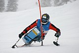 Para-Alpinskifahrer in voller Action auf der Skipiste, konzentriert und dynamisch mit Krückenski und blauer Rennkleidung. Schneebedeckte Landschaft im Hintergrund.