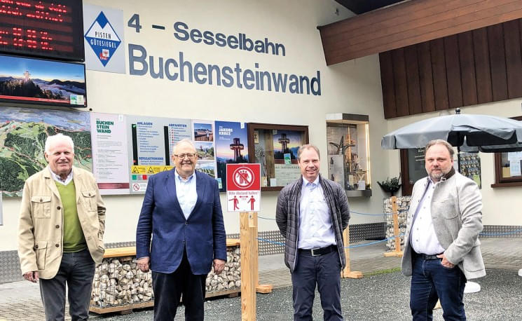 Vier Männer stehen vor dem Eingang einer Sesselbahn mit dem Namen 'Buchensteinwand', umgeben von Infotafeln und einer Sitzbank aus Holz.