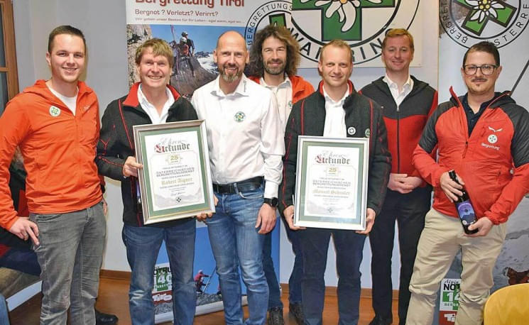 Gruppe von sieben Männern in Freizeitkleidung, zwei halten Urkunden. Sie stehen vor einem Banner mit einem Logo. Alle lächeln und tragen Jacken.