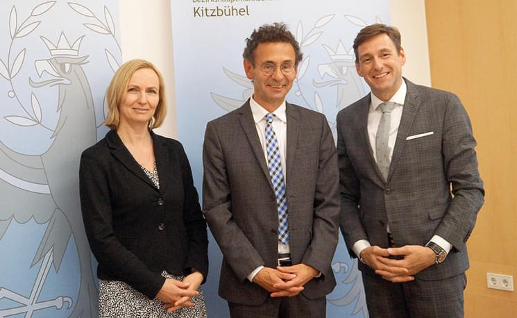 Drei Personen posieren in Businesskleidung vor einem Banner mit einem Wappenmotiv. Sie lächeln in die Kamera und stehen in einem gut beleuchteten Raum.