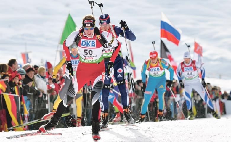 Biathlon-Wettbewerb mit mehreren Athleten in Aktion; sie laufen auf schneebedeckter Strecke und tragen Skiausrüstung, umgeben von Zuschauern und Flaggen.