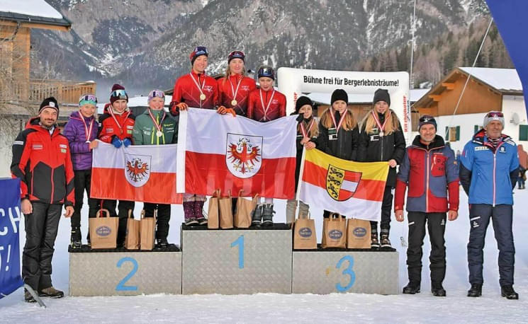 Drei Teams stehen auf einem Podest bei einer Siegerehrung im Freien, umgeben von Bergen, und halten Fahnen sowie Medaillen. Ein winterliches, sportliches Event.