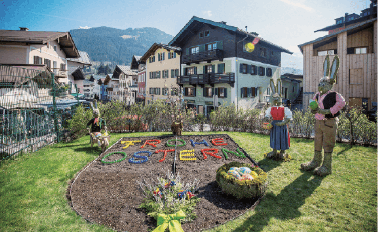 Ein bunt dekorierter Ostergruß in einem Garten mit Schriftzug "Frohe Ostern" aus Blumen, umgeben von lebensgroßen Hasenfiguren, in einem alpinen Dorf.