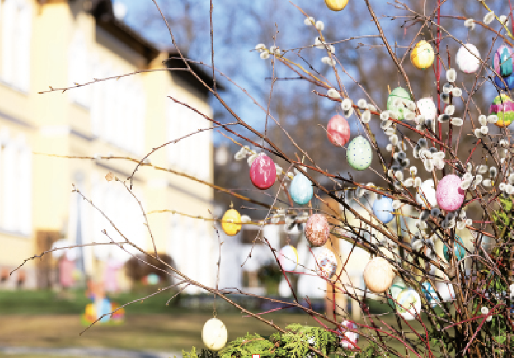 Ein blühender Strauch, geschmückt mit bunten Ostereiern, steht im Vordergrund. Im Hintergrund sind verschwommene Gebäude und blauer Himmel sichtbar.