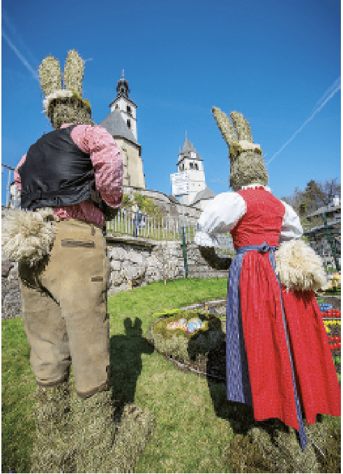 Zwei Personen in Hasenkostümen stehen vor einer Kirche mit einem Korb voller bunter Eier. Die Szene wirkt frühlingshaft und festlich.