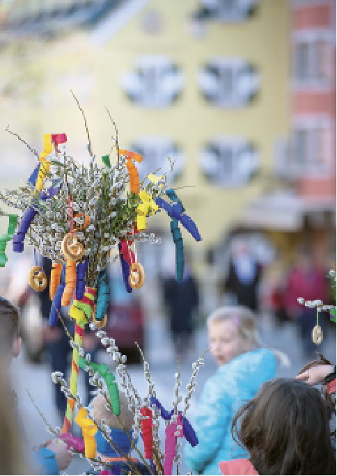 Ein dekorativer Zweig mit buntem Band umwickelten Ästen und getrockneten Orangenscheiben, unscharfer Hintergrund mit Menschen und Gebäuden im Freien.