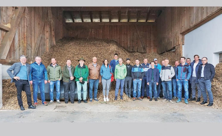 Eine Gruppe von Menschen steht nebeneinander in einem Holzlager, gefüllt mit Holzspänen, alle lächeln in die Kamera. Es sind sowohl Männer als auch Frauen dabei.