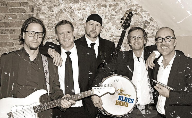 Fünf Männer in Anzügen posieren mit Musikinstrumenten, darunter eine Gitarre und ein Schlagzeug mit dem Logo "Legacy Blues Band" auf der Bassdrum.