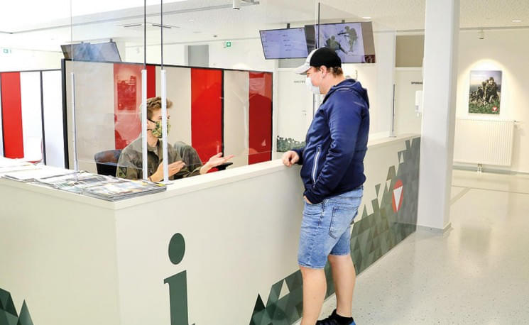 Ein Mann steht an einem Empfangstresen in einem modernen Büro, spricht mit einer Person dahinter. Bildschirme und rote Trennwände im Hintergrund.