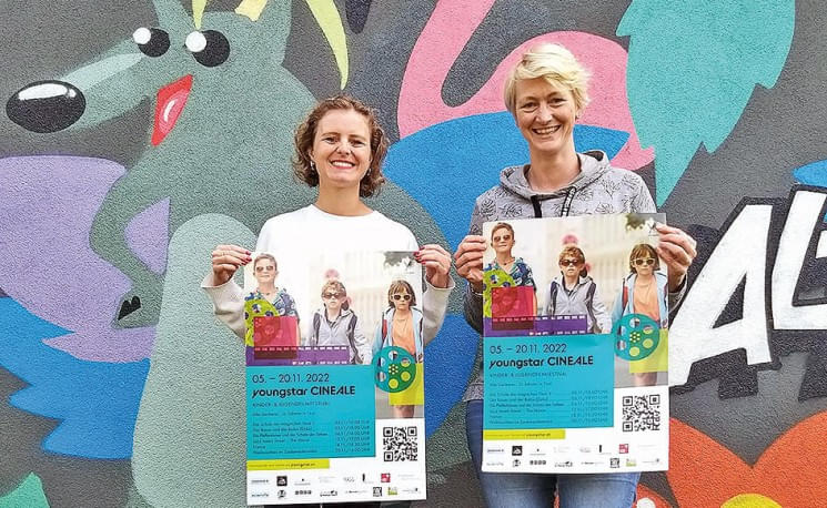 Zwei Frauen posieren vor einer bunten Wandmalerei und halten jeweils Plakate in den Händen. Die Plakate werben für eine Veranstaltung mit musikbezogenem Thema.