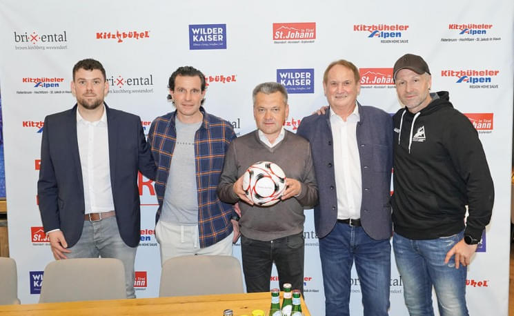 Fünf Männer stehen nebeneinander vor Sponsoren-Logos, der mittlere hält einen Fußball. Sie tragen lässige Kleidung und lächeln in die Kamera.